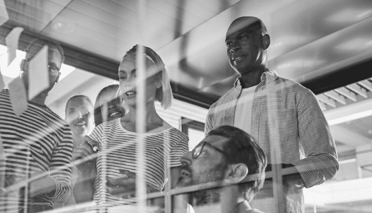 A black and white photo of a group of professionals collaborating and brainstorming by writing on a clear glass wall in an office.
