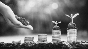 A black and white photo of a hand adding soil to sprouts growing on a stack of coins.