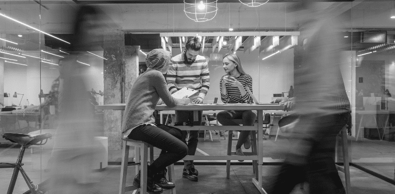 A black and white, blurry photo of a group of people working together in a modern, glass-walled office.