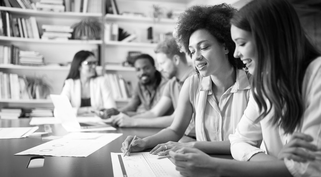A black and white photo of a diverse group of people collaborating at a table. Two women in the foreground are smiling and looking at documents.