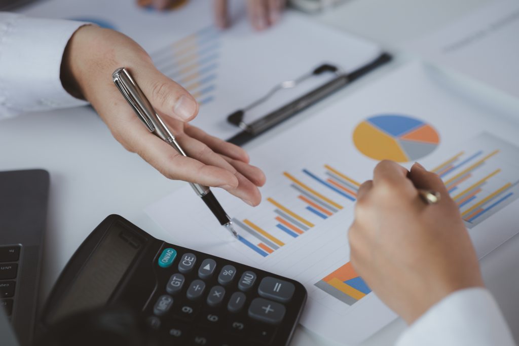 A close-up of two people's hands pointing at various graphs, charts, and a calculator on a desk.